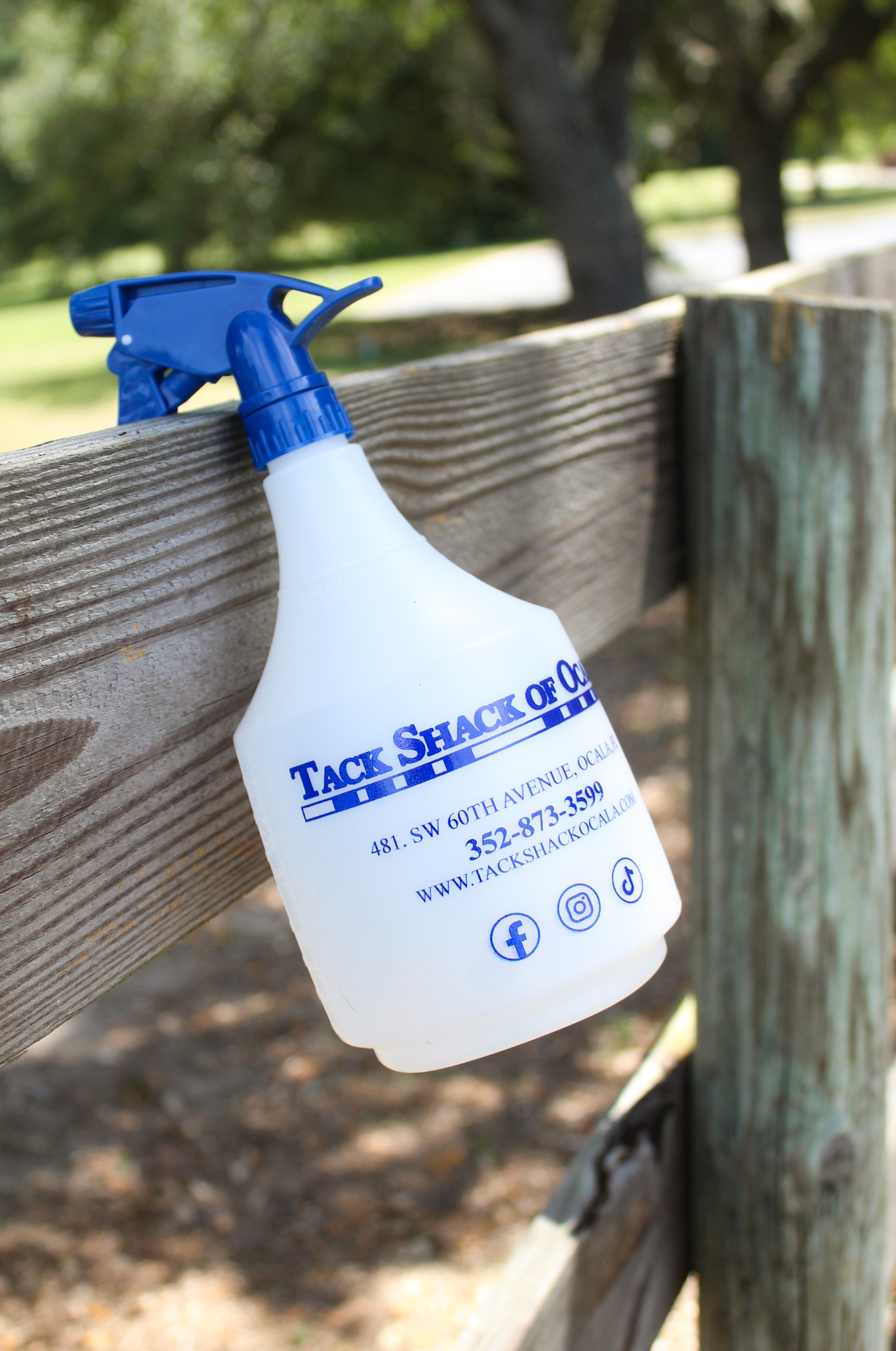 A white spray bottle with blue text and a blue trigger spray head. The text includes the name 'TACK SHACK OF OCALA' and contact information.