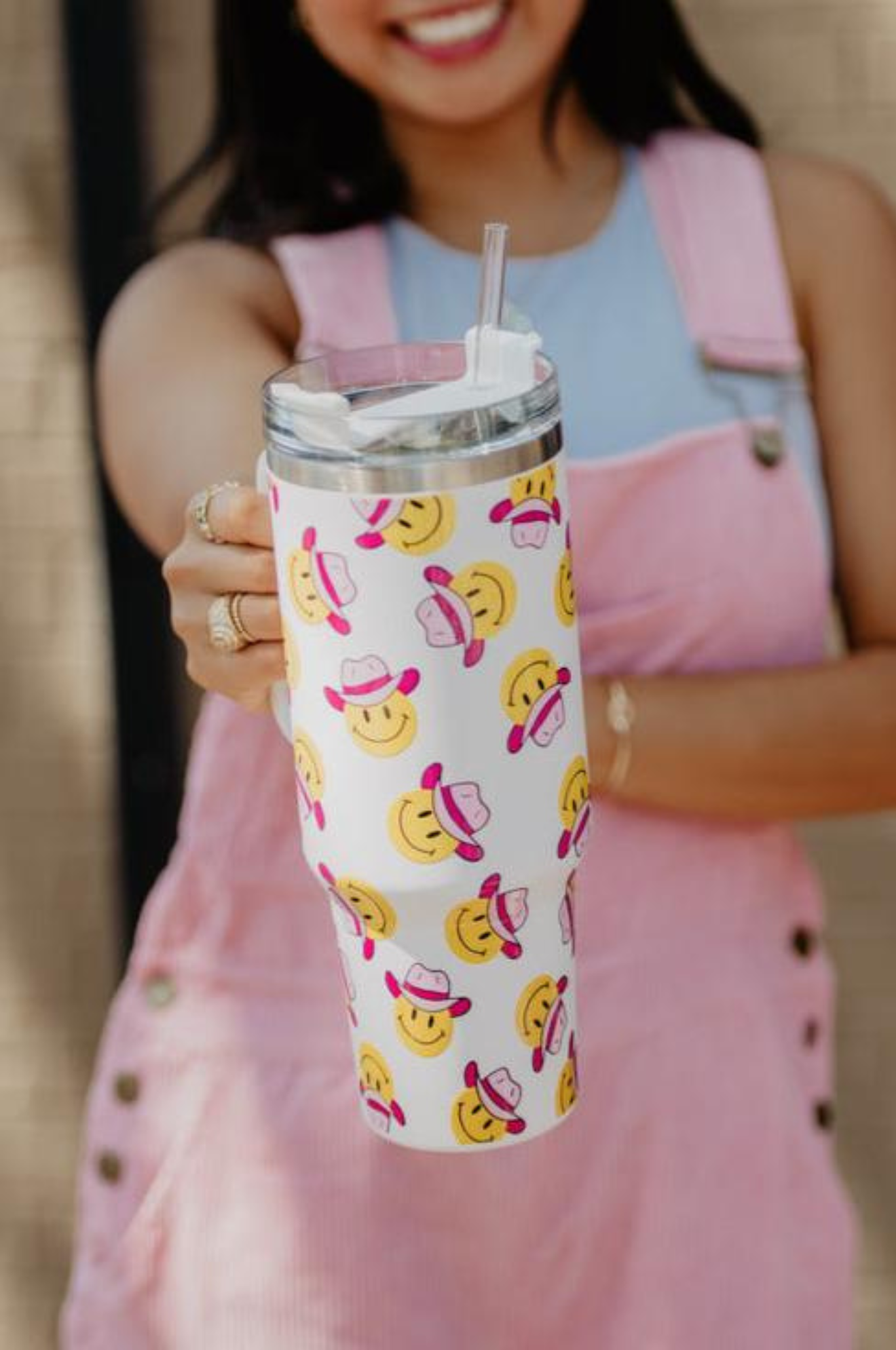 A white stainless steel tumbler with a handle, featuring a repeated pattern of happy faces wearing pink cowboy hats.
