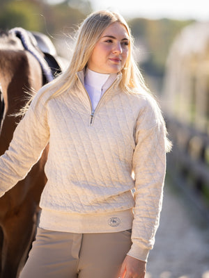 Woman wearing a beige quilted jacket standing next to a horse in an outdoor setting