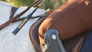 A close-up of a horse bit with a black handle and yellow 'The Shire of Texas' logo, resting on brown leather riding boots and a brown leather saddle on a white horse.
