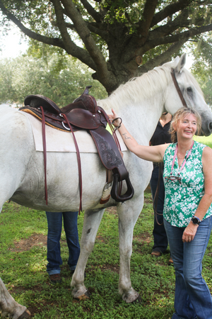 A woman standing next to a white horse fitted with a brown saddle, with trees and sky in the background.