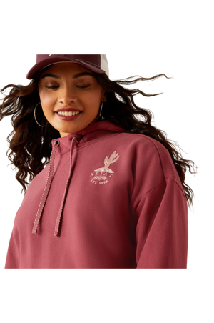 A female model wearing a maroon hoodie with a logo on the front, paired with blue denim shorts and a matching maroon cap.