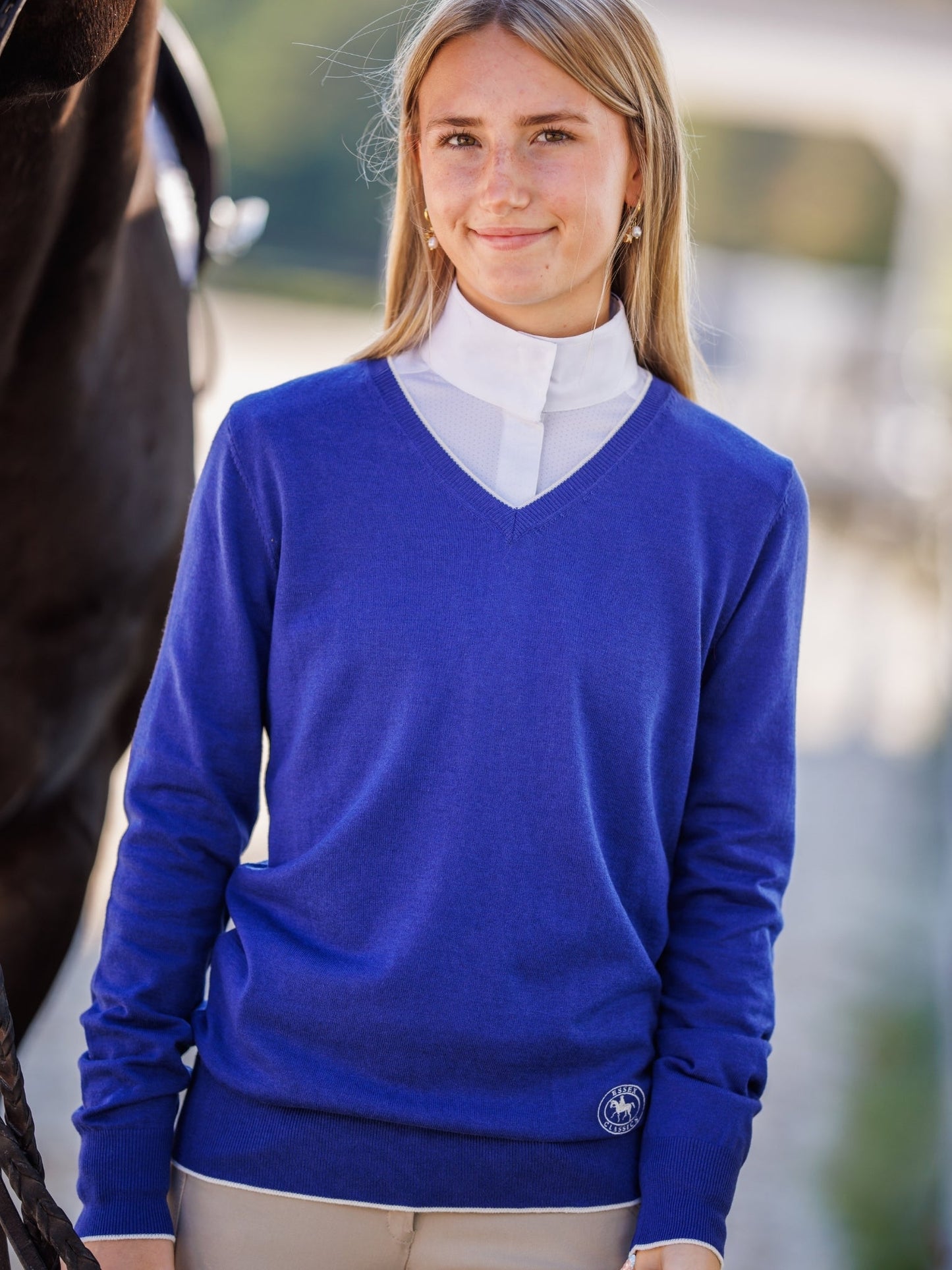 Woman wearing a blue sweater with a white shirt underneath, standing next to a horse.