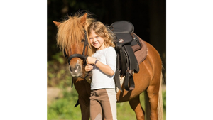 Young girl standing next to a brown horse with a saddle in a natural setting
