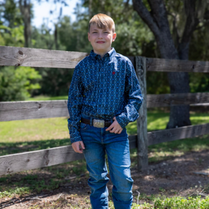 Young boy in a blue patterned shirt and jeans standing in front of a wooden fence.
