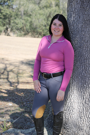 Person wearing a pink top and gray riding pants leaning against a tree outdoors.