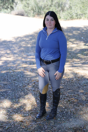Person wearing equestrian attire including a blue top, gray riding pants, and black boots outdoors.