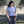 Woman wearing a light purple shirt and gray pants standing outdoors with trees in the background