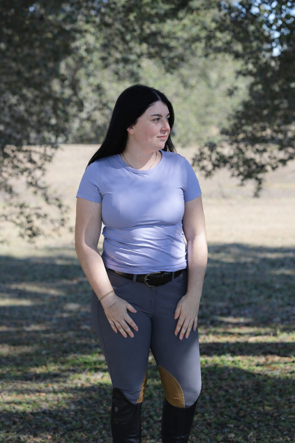Woman wearing a light purple shirt and gray pants standing outdoors with trees in the background