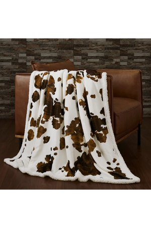 A sherpa throw with a cowhide print pattern in beige, brown, and white colors.