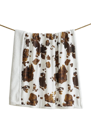 A sherpa throw with a cowhide print pattern in beige, brown, and white colors.