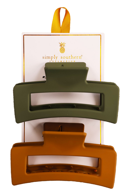 Two hair clips, one green and one brown, displayed on a vertical cardboard stand with a logo at the top.