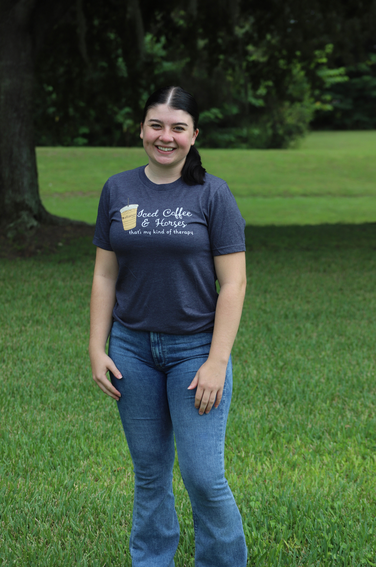 Person wearing a blue t-shirt with text and graphics, standing in a grassy area with trees.