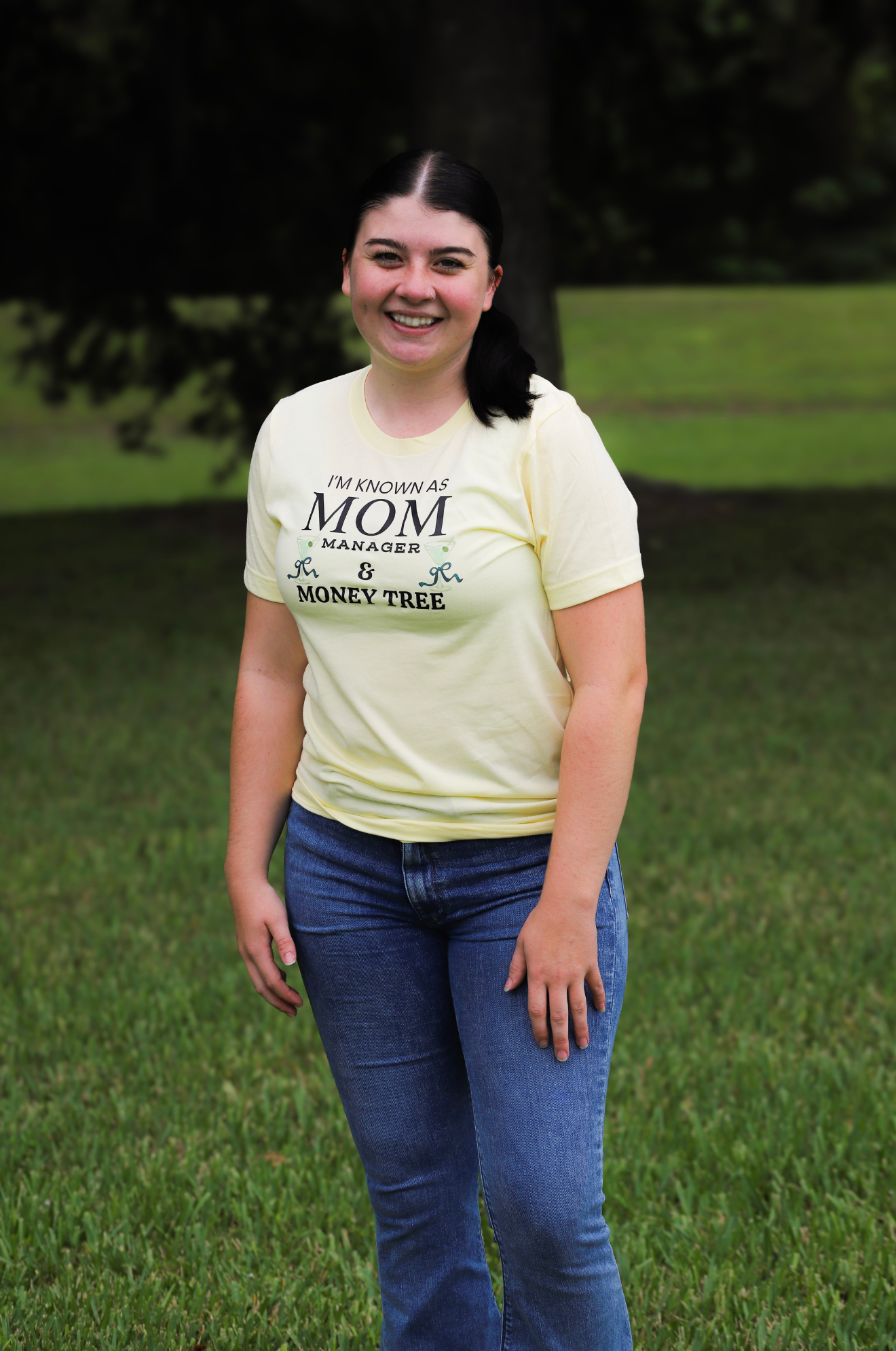 Person wearing a light yellow t-shirt with text standing in a grassy area.