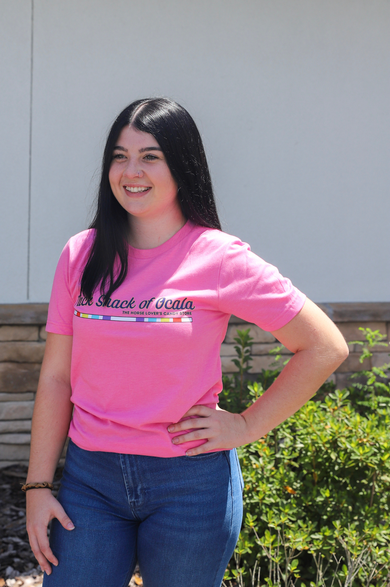 Woman wearing a pink t-shirt with text, standing outdoors against a white wall.