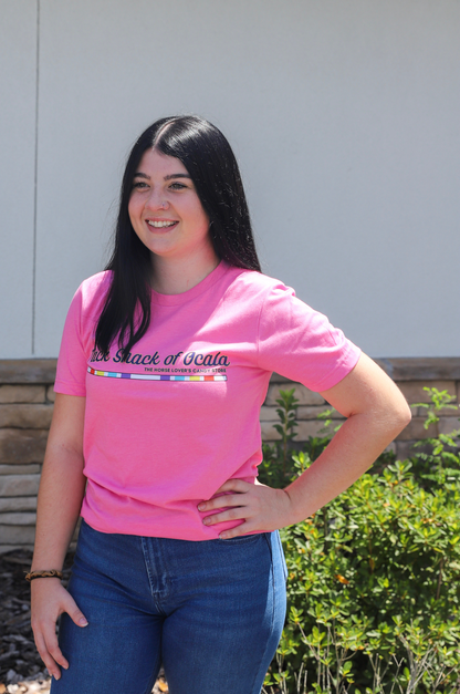 Woman wearing a pink t-shirt with text, standing outdoors against a white wall.