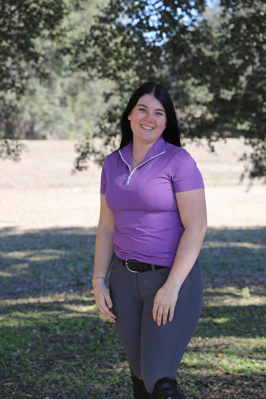 Woman wearing a purple shirt and gray pants standing outdoors with trees in the background